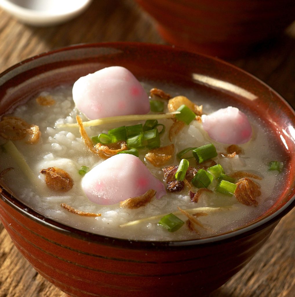 A bowl of congee topped with chili-flavored fish balls, garnished with green onions and sesame seeds.